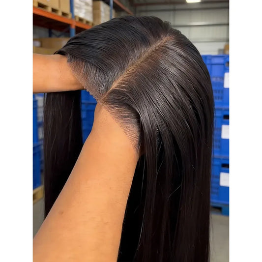 A close-up of a person holding a long, straight, dark brown to black hair extension or wig piece, showing the hair part and texture. The background shows a warehouse setting with blue storage bins and shelving.